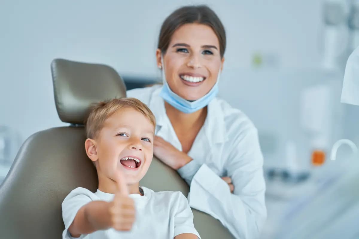 Menino sorridente fazendo sinal de positivo na cadeira do dentista, ao lado da dentista