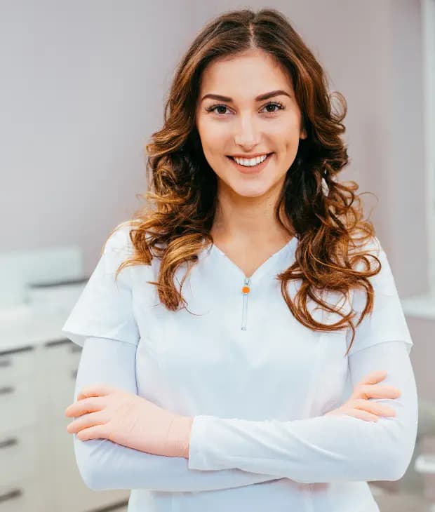 Dentista de uniforme branco sorrindo, com os braços cruzados, em um consultório odontológico