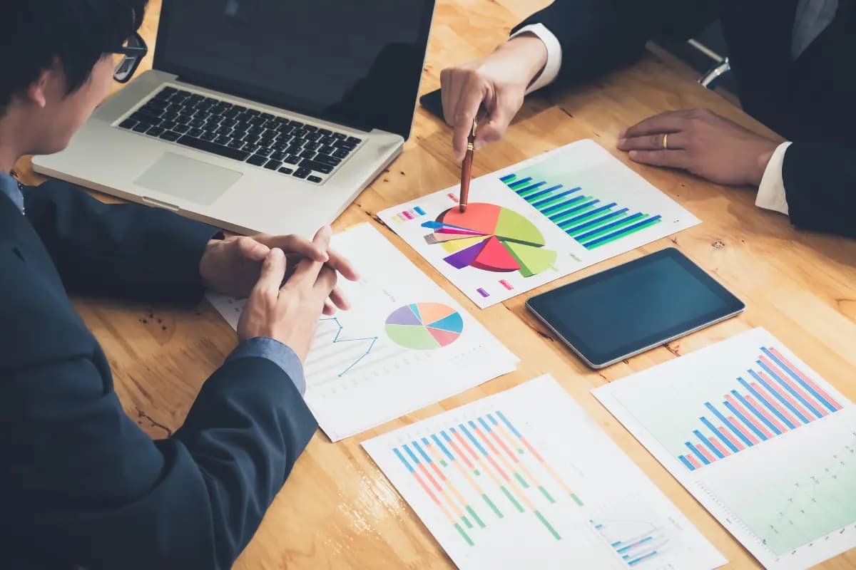 Duas pessoas analisam gráficos e relatórios impressos sobre a mesa, comparando opções com notebook e tablet